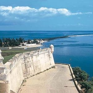 Una cinta muraria in pietra domina un mare blu calmo e una penisola sabbiosa con alberi sotto un cielo parzialmente nuvoloso.