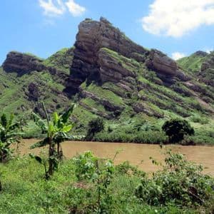 Un fiume marrone fangoso scorre attraverso una valle verde e lussureggiante con banani, sullo sfondo di grandi montagne rocciose sotto un cielo blu.
