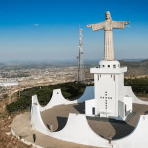 Una veduta aerea di una grande statua di Cristo, posta su una piattaforma bianca in cima a una collina, che domina un'ampia città sotto un cielo blu.