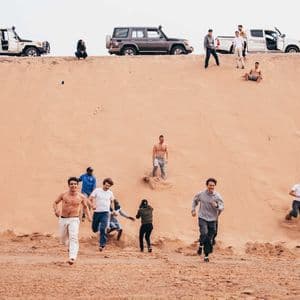 Un viaggio di gruppo WeRoad con persone che corrono e giocano su una grande duna di sabbia, con veicoli 4x4 parcheggiati sulla cresta.