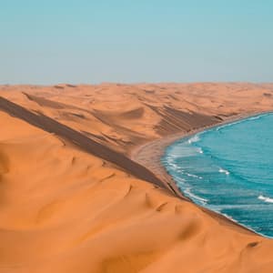 Vaste dune di sabbia arancione incontrano l'oceano blu lungo una costa sotto un cielo sereno.