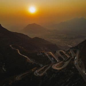 Veduta aerea di una strada tortuosa con tornanti che scende da una montagna, con auto che la percorrono durante un tramonto velato.