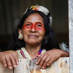 Una anciana indígena con pintura facial roja y un tocado de plumas mira a través de un marco de ventana de madera.