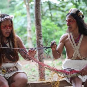 Deux femmes autochtones en tenue traditionnelle et coiffes à plumes travaillent ensemble avec des fils colorés en extérieur.