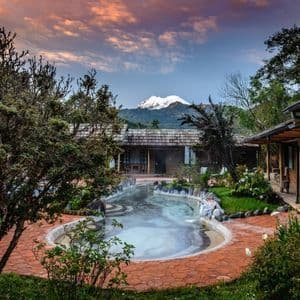 Una piscina termale fumante in un resort con chalet in legno, sullo sfondo di una montagna innevata sotto un cielo al tramonto.
