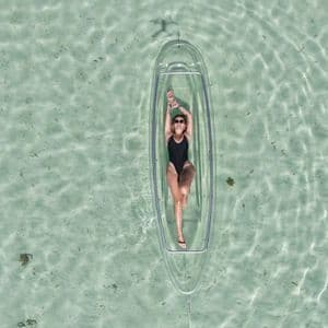 Vista dall'alto di una donna in costume nero sdraiata in un kayak trasparente che galleggia su acqua limpida e poco profonda.
