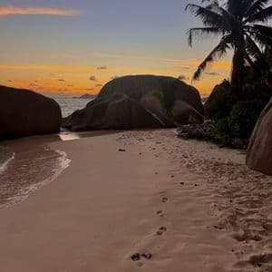 Impronte nella sabbia su una spiaggia tropicale con grandi massi e una palma durante un tramonto colorato.