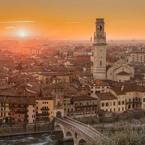 Una veduta aerea di una città storica con tetti in terracotta, un ponte di pietra su un fiume e un imponente campanile al tramonto.