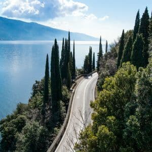 Veduta aerea di una strada tortuosa in riva al lago circondata da cipressi, con montagne visibili al di là dell'acqua.