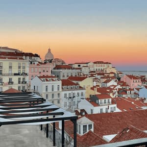 A cityscape of colorful buildings with red-tiled roofs seen from a terrace at sunset, with the sea in the distance.