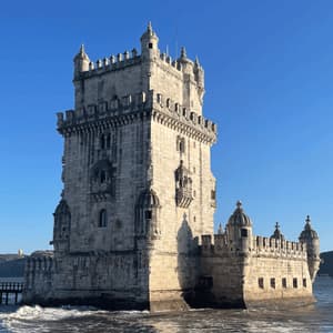 An ornate stone fortress with turrets stands in the water next to the shore under a clear blue sky.