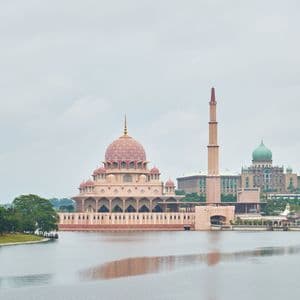 Une grande mosquée aux dômes roses ouvragés et à un haut minaret se dresse au bord d'une étendue d'eau sous un ciel nuageux.