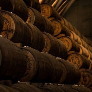 Botti di legno scuro allineate in file ordinate all'interno di una cantina di distilleria in penombra, con soffitto ad arco.