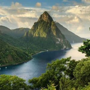 Una grande montagna conica verde sulla costa di una baia blu, con un piccolo villaggio e barche a vela sull'acqua.
