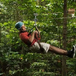 Un uomo con casco e imbracatura blu sorride mentre scende in zipline su un cavo attraverso una fitta giungla verde.