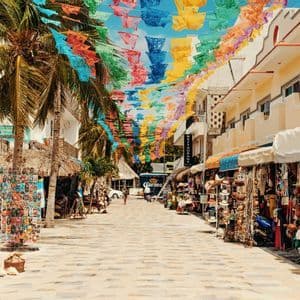 Striscioni decorativi colorati sono appesi lungo una strada pedonale soleggiata, fiancheggiata da palme e bancarelle di souvenir.