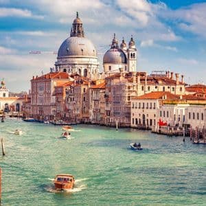 Un ampio canale con diverse barche è fiancheggiato da edifici storici, con una grande basilica a cupola sullo sfondo sotto un cielo azzurro e nuvoloso.