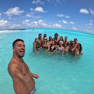 Ein Mann macht ein Selfie von einer lächelnden WeRoad-Gruppe, die gemeinsam im türkisfarbenen Meerwasser nahe einem weißen Sandstrand steht.