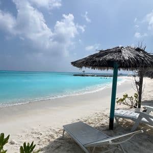 Liegestühle und Strohschirme an einem weißen Sandstrand blicken auf ruhiges, türkisfarbenes Wasser unter einem teilweise bewölkten Himmel.