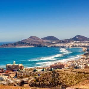 Una vista panoramica di una città costiera lungo una spiaggia sabbiosa, con un oceano turchese e montagne aride sotto un cielo azzurro e limpido.