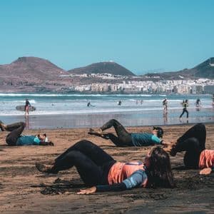 Un viaggio di gruppo WeRoad di persone in muta che fanno stretching su una spiaggia sabbiosa, con surfisti nell'oceano e una città sullo sfondo.