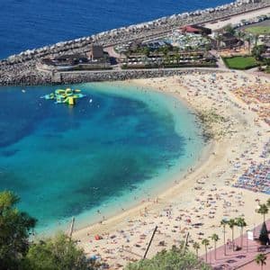 Veduta aerea di una spiaggia sabbiosa affollata a forma di mezzaluna, con persone che prendono il sole sotto ombrelloni colorati e nuotano in acque turchesi.