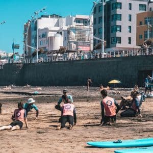 Un viaggio di gruppo WeRoad si prepara su una spiaggia sabbiosa per una lezione di surf, con tavole da surf in primo piano e una città sullo sfondo.