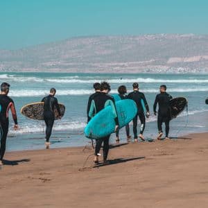 Un viaggio di gruppo WeRoad con persone in muta che trasportano tavole da surf e camminano verso l'oceano su una spiaggia sabbiosa.
