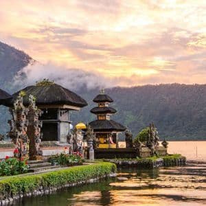 Un temple orné à plusieurs niveaux se dresse sur la rive d'un lac calme au coucher du soleil, avec des montagnes boisées en arrière-plan.