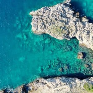 An aerial top-down view of a rugged, light-colored rocky shoreline meeting a clear, turquoise sea.
