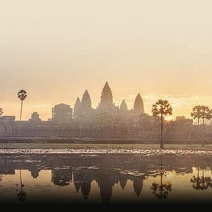 The Angkor Wat temple silhouetted against a golden sunrise, its form reflected in a tranquil pond surrounded by palm trees.
