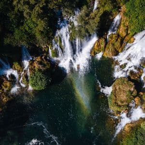 Vista aerea dall'alto di cascate che si gettano su rocce muschiose e alberi in una pozza turchese, con un arcobaleno nella nebbia.
