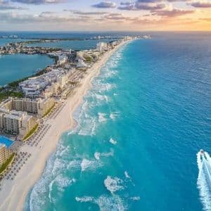 Veduta aerea di una costa con hotel lungo una spiaggia di sabbia bianca bagnata da acque turchesi, mentre un motoscafo sfreccia al tramonto.