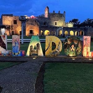 Lettere illuminate e colorate che compongono 'VALLADOLID' su un prato verde, con un vecchio edificio in pietra illuminato sullo sfondo al tramonto.