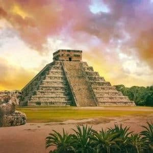 Un'imponente piramide Maya a gradoni in un sito archeologico, circondata da rovine di pietra e vegetazione sotto un cielo colorato al tramonto.