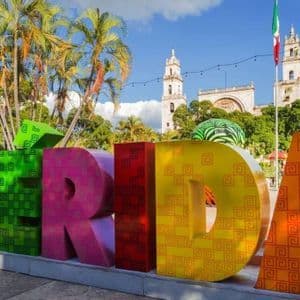 Grandi lettere colorate che compongono la parola 'MERIDA' si ergono in una piazza con una cattedrale, palme e una bandiera messicana sullo sfondo.