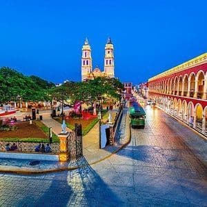 Una piazza cittadina al crepuscolo con una grande cattedrale sullo sfondo, un edificio rosso con archi e un tram verde su una strada di ciottoli.