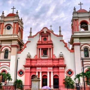 Symmetrische Fassade einer reich verzierten Kirche mit rosa-weißen Mauern, zwei Glockentürmen und Kreuzen vor bewölktem Himmel.