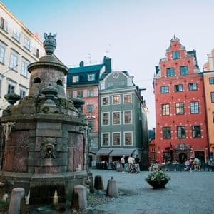An ornate stone fountain stands in a cobblestone square, with colorful historic buildings in the background under a clear sky.