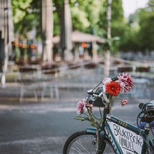 A close-up of a bicycle's handlebars decorated with red and pink flowers, with a blurry outdoor cafe in the background.