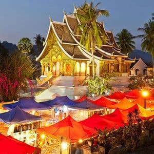 Tende colorate rosse e blu di un mercato notturno brillano davanti a un tempio dorato illuminato al tramonto.