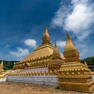 Una grande stupa dorata e riccamente decorata, con molteplici guglie, si erge in un cortile pavimentato sotto un cielo azzurro con nuvole.
