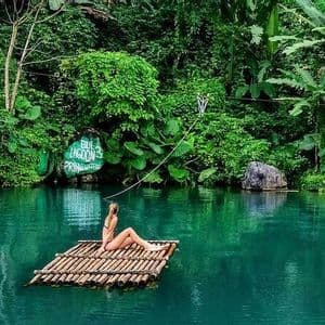 Una donna in bikini siede su una zattera di bambù che galleggia sulle acque calme e verdi di una laguna circondata da una giungla lussureggiante.