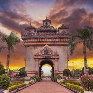 Al termine di un sentiero, in un parco con palme, un grande e ornato monumento ad arco si staglia contro un cielo colorato al tramonto.
