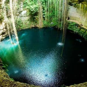 Una veduta grandangolare dall'alto di un grande cenote a cielo aperto con acqua blu profonda, circondato da pareti rocciose e piante rampicanti pendenti.