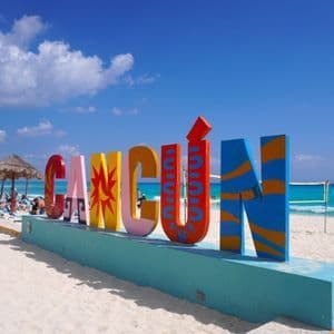 Un grande cartello colorato con la scritta 'Cancún' si trova su una spiaggia di sabbia bianca accanto all'oceano turchese sotto un cielo azzurro brillante.
