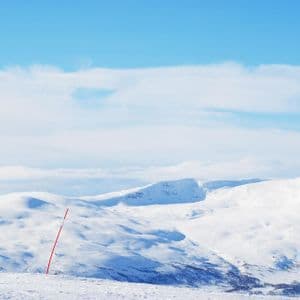 Un segnavia sulla pista da sci si staglia su un pendio innevato, affacciato su una vasta catena montuosa coperta di neve sotto un cielo azzurro.