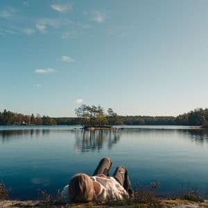 A person lies on a rocky shore, looking out over a calm lake with a small, tree-covered island in the distance.