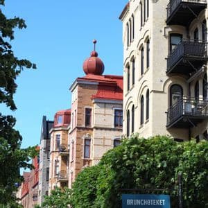 Ornate European buildings with a red turret stand along a street with leafy green trees and a sign reading 'BRUNCHOTEKET'.