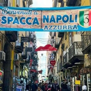 A blue banner reading 'Spacca Napoli' hangs over a crowded, narrow street lined with shops and yellow buildings.